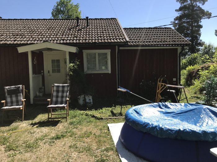 Rödfärgat hus med variabel takhöjd, ytterdörr till höger och en uppblåsbar pool framför, samt trädgårdsmöbler på gräset.