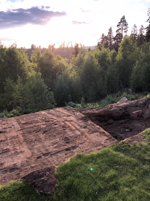 Utgrävt område för pool i en slänt med skymd solnedgång och träd i bakgrunden.