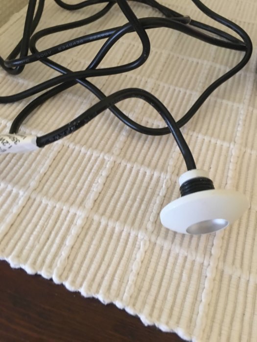 Vit kabelgenomföring med medföljande clips och svart elektrisk kabel på ett beige mattunderlag.