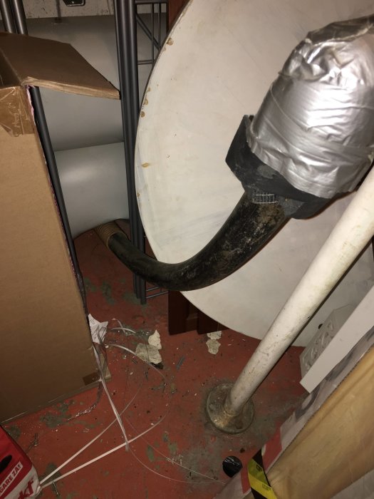 Svart slang ansluten till vägg med tejp, omringad av rör och bråte på ett stökigt golv.