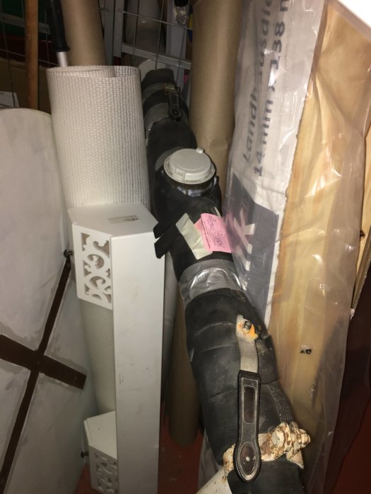 Svart rörledning med två reglage och en slang som går in i en vägg i en källare, omgiven av orelaterade föremål.