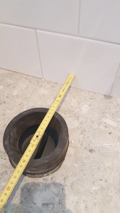 Måttband som mäter avståndet mellan vägg och avloppsrör på toalettgolv.