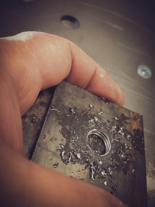 Närbild av en hand som håller ett metallstycke med nyligen borrade 10.2mm hål och metallspån.