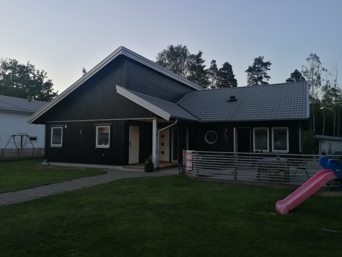 Svart funkisstil hus med grå takpannor, vit dörr med rutor och röda lister, och leksaker på gräsmattan.