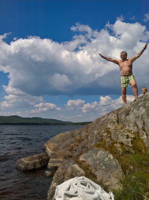 Person med armarna utsträckta på en klippa vid en sjö under en solig dag.