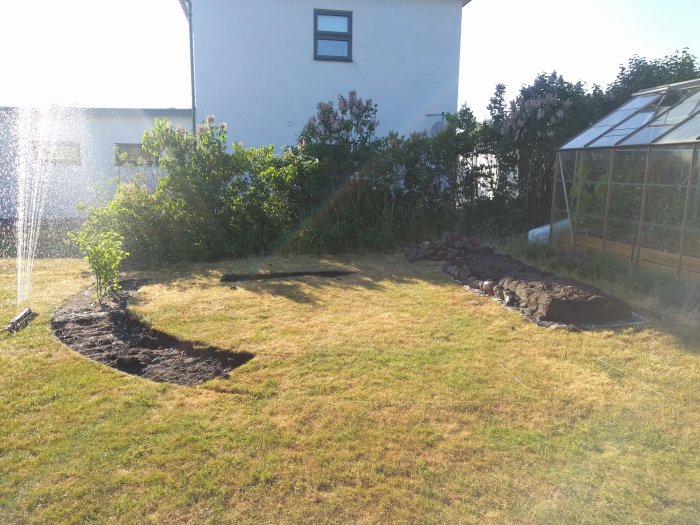Trädgård under arbete med en halvfärdig jordgubbsås bestående av grässvål och jord anlagd längs en stenrad, intill ett växthus.