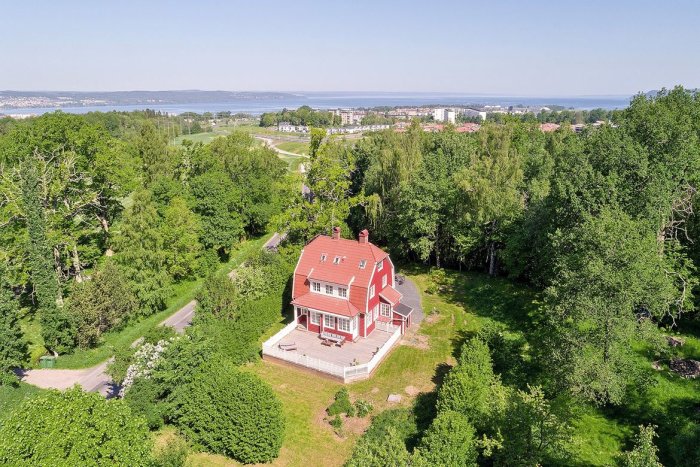 Röd villa omgiven av grönska med utsikt över stad och vatten i bakgrunden.