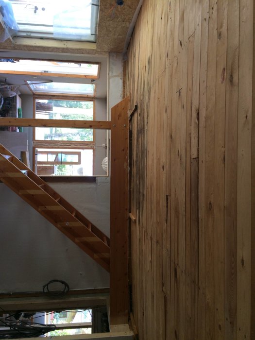 Ett vardagsrum under renovering med en trävägg och trappa, och synliga takstolar som hålls upp av en stolpe.