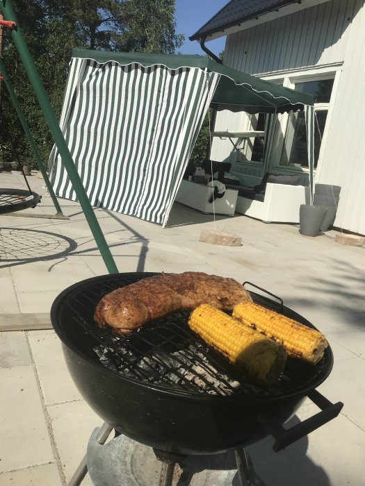 En grill med stekande köttbit och majs på en solig uteplats med vit husfasad och gröna markiser i bakgrunden.