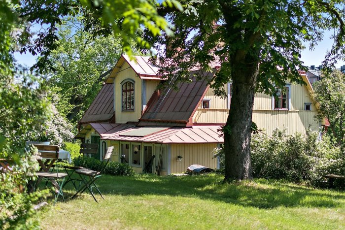 Gult 1700-talshus med tegeltak och glasveranda omgivet av en grönskande trädgård och terrassering på Långholmen, Stockholm.