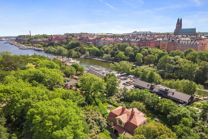 Flygbild av Ingenjörsbostaden omgiven av grönska på Långholmen i Stockholm, med stadsutsikt och vatten.