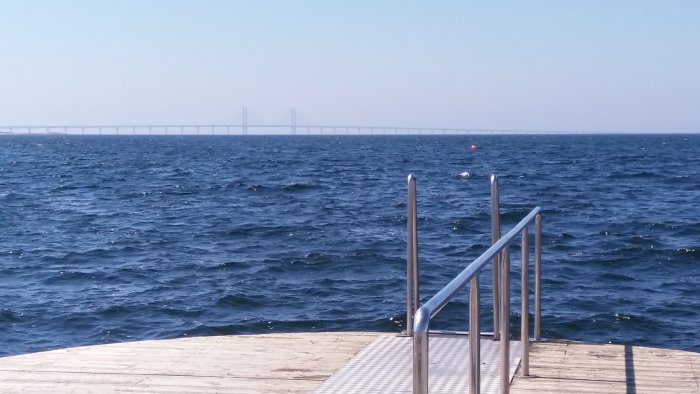 Vy över havet från en brygga med räcke, bro i bakgrunden under klar himmel.