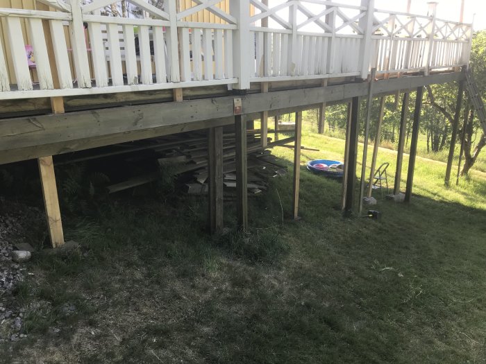 Träaltan byggd på pålar med vit räcke och en gräsmatta nedanför, lite byggmaterial synligt under altanen.