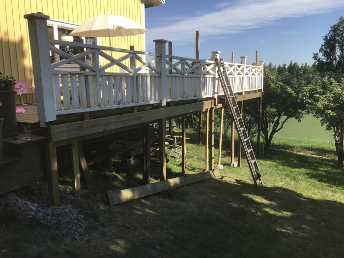 Byggd träaltan med räcke och stege utomhus vid ett gult hus omgivet av grönska.