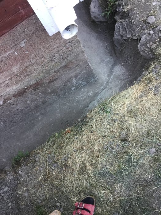 Grävd dräneringsgrop vid källarmur på 1930-tals hus, synlig berggrund och avloppsrör, samt röd sandal i förgrunden.