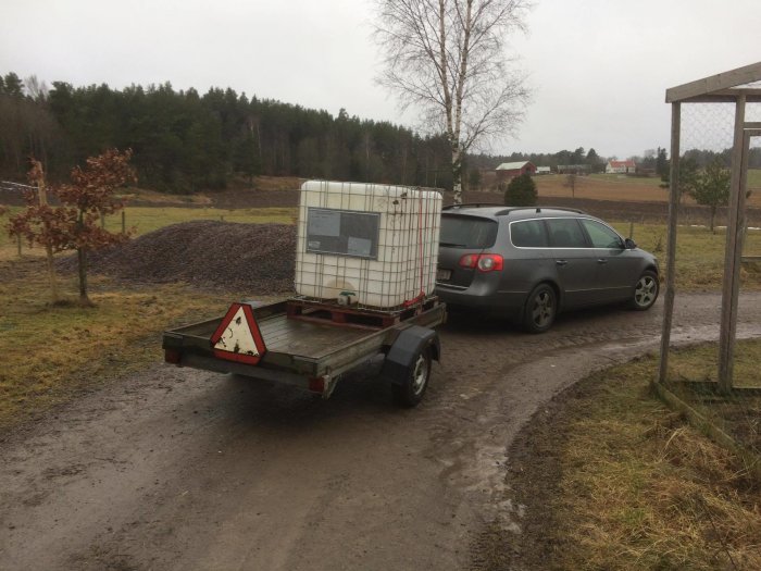 IBC plasttank monterad på en släpkärra, parkerad vid en grusväg med landskap i bakgrunden.