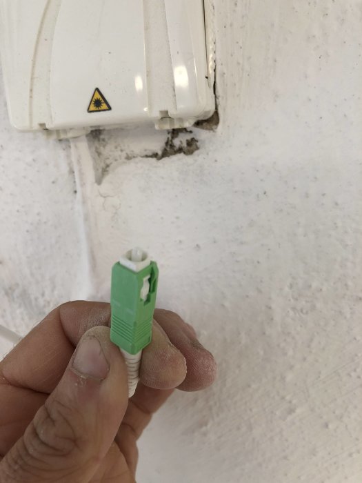 Hand håller en trasig grön RJ45 Ethernet-kontakt framför en nätverksuttag i väggen.