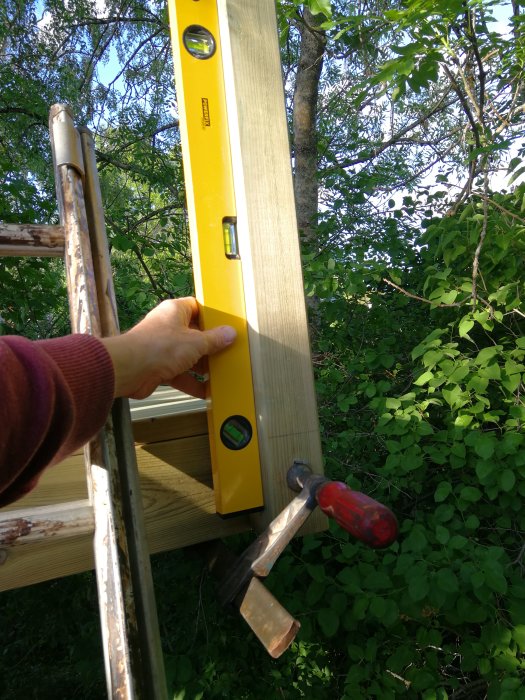 Hand håller ett vattenpass mot en trästolpe med en hammare och verktyg i förgrunden, naturbakgrund.