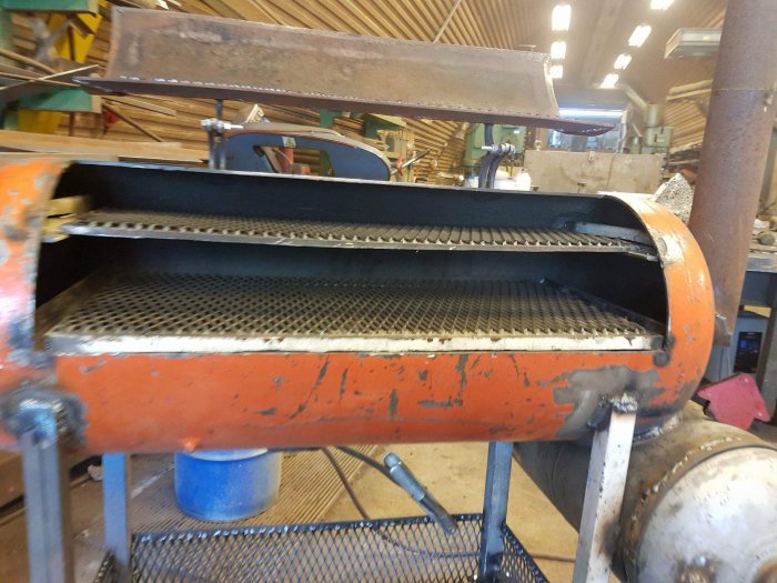 Sliten, orange byggmaskin med uppfälld lock och synlig metallgaller, i ett verkstadsutrymme.