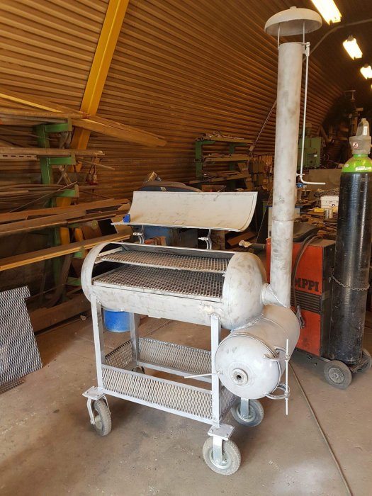 En hemmagjord mobilt vedeldad grill med smutsig yta i ett verkstadsutrymme med verktyg och trämaterial i bakgrunden.