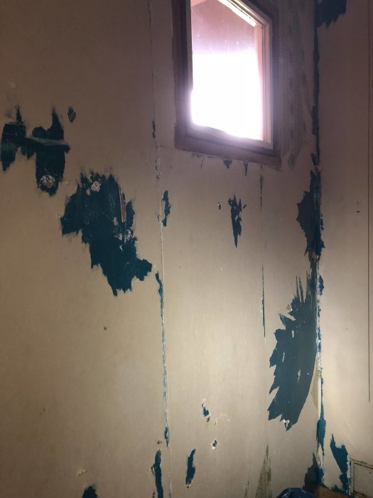 En vägg i ett rum med masonitpaneler där gammal tapet delvis är bortskrapad och spikhål syns.