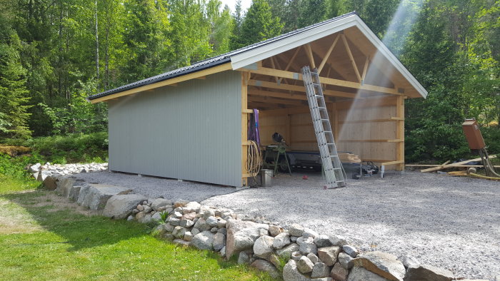 Nybyggd trädgårdsbod med grå panel på ena långsidan och synlig träkonstruktion på gaveln, omgiven av natur och grusgång.