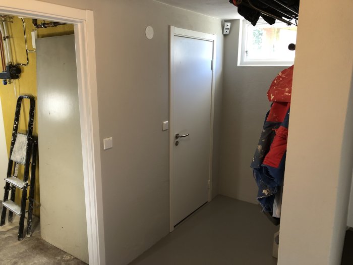 Högblankt grått golv och ljusgråa väggar i ett renoverat rum med vit dörr och foder, stege och överdragen stege syns.