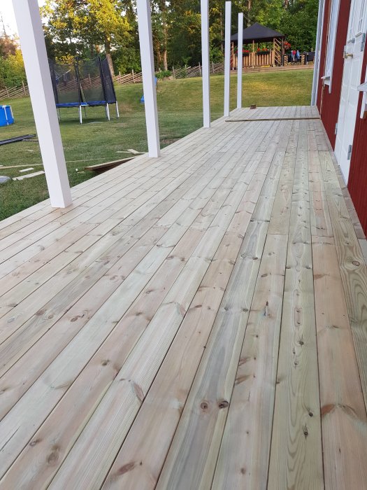 Nybyggd veranda med trallgolv, nästan färdigt med omonterade skruvar synliga, och en trädgård i bakgrunden.