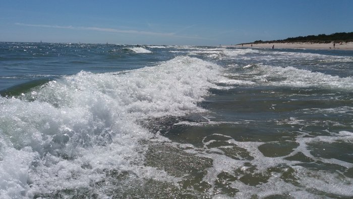 Vågor som bryts nära kusten på en solig dag med strand och människor i bakgrunden.
