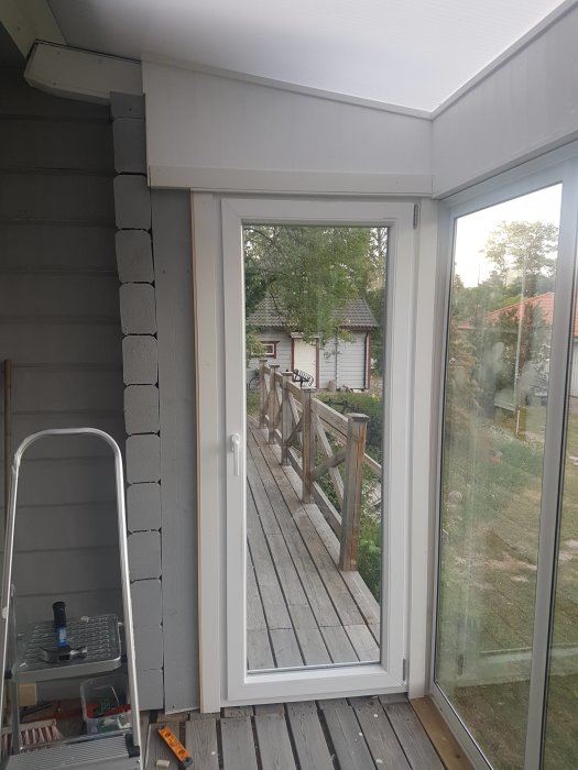 Nyinstallerad vit dörr i sned husvägg med specialanpassat dörrfoder på en inglasad veranda, verktyg syns på golvet.