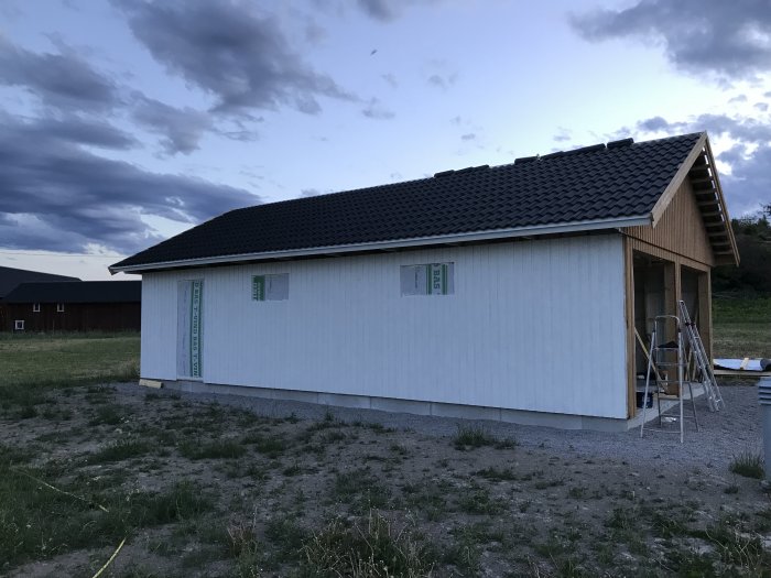 Ett halvfärdigt hus i skymningen med en stege mot fasaden under pågående målningsarbete.