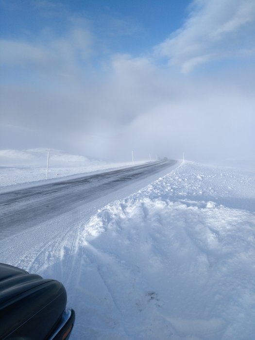 Vinterväg över fjällandskap med snö och dimma, bilens sidospegel syns i förgrunden.