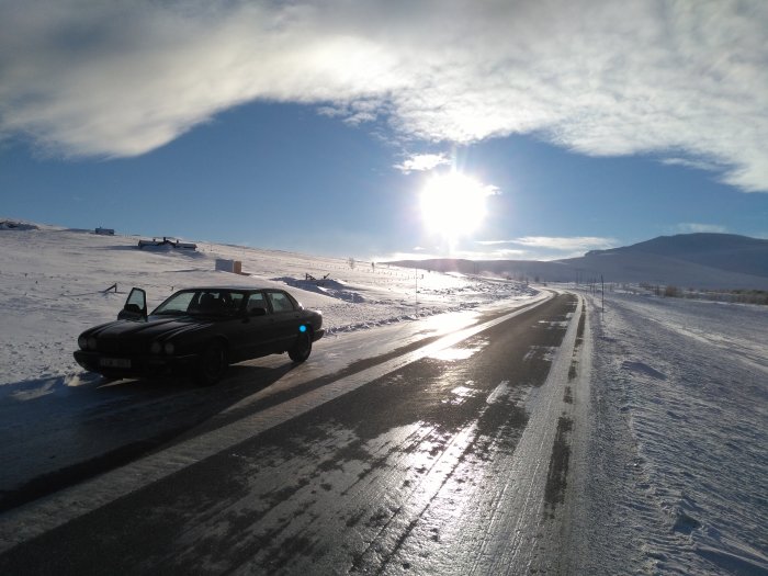 Bil parkerad på snöig väg med öppen dörr mot solig fjällvy.