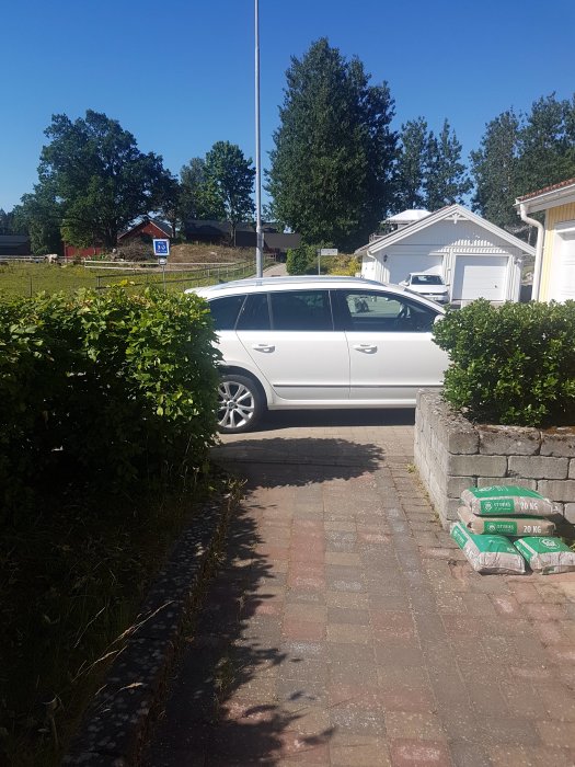 Vit personbil parkerad bredvid häck intill uppfart av stenplattor och säckar med byggmaterial.