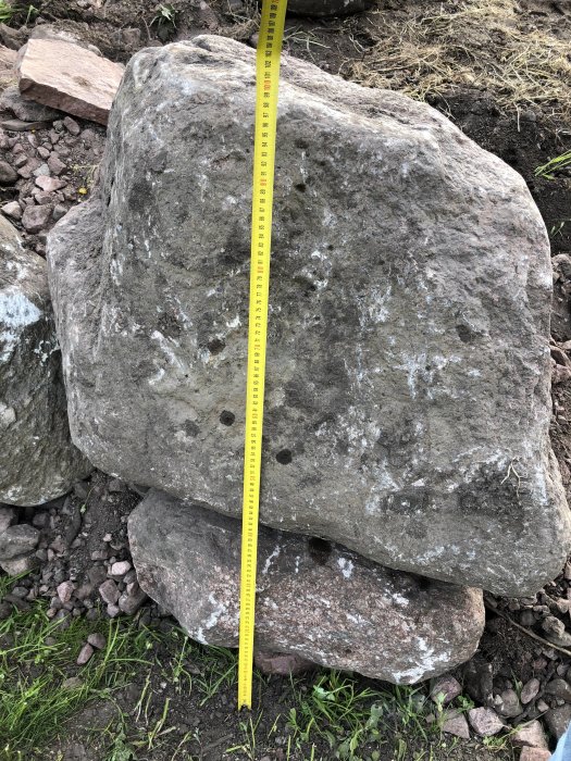 Stor sten använd för stödmur med måttband som visar höjden, står på grus och mindre sten.