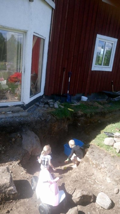 Två barn leker vid grunden av ett rött hus under renovering med leksaksbilar.