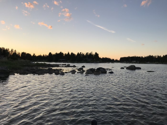 Solnedgång över en vattensamling med stenar och en strandlinje med träd i bakgrunden.