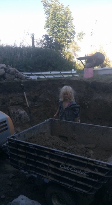 Barn står i en skottkärra framför en grävd grop för poolbygge med byggverktyg och rep i bakgrunden.