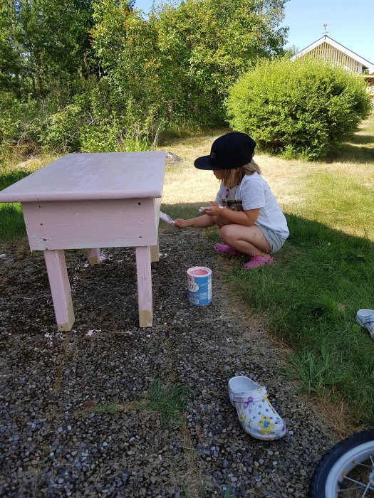 Barn målar en rosa arbetsbänk av trä utomhus, med öppen färgburk och pensel i handen.