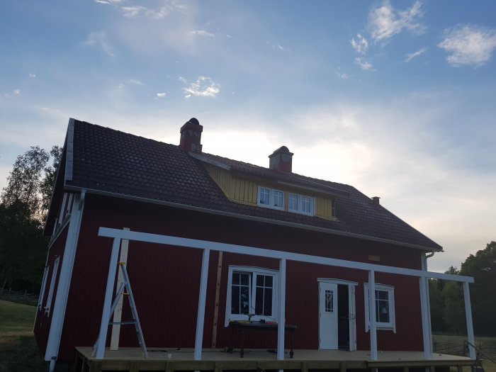 Rödmålat hus med nyinstallerad främre balk och pågående takarbeten mot en aftonhimmel.