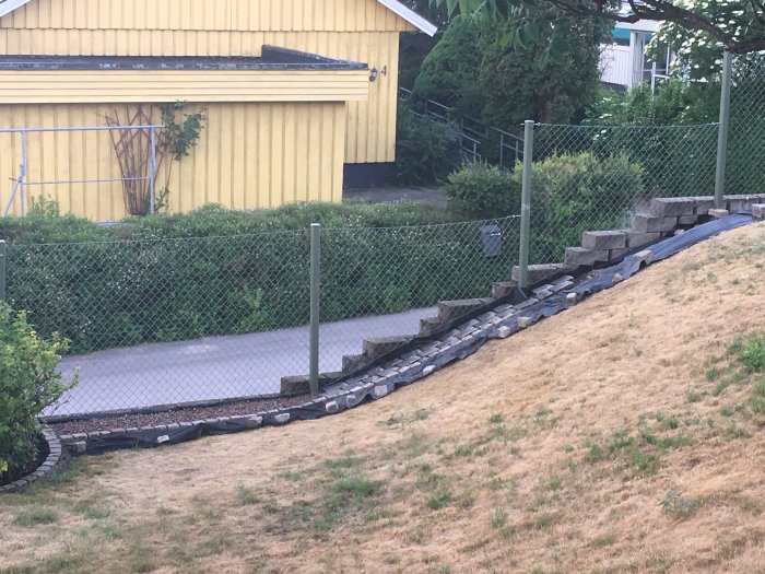 Stenlagd kant längs sluttning med torkad gräsmatta framför gult hus och staket.