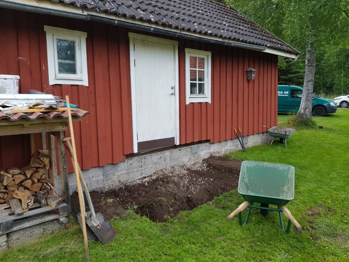 Nybyggd trätrappa vid röd uthusdörr med pågående markarbete och skottkärra framför.