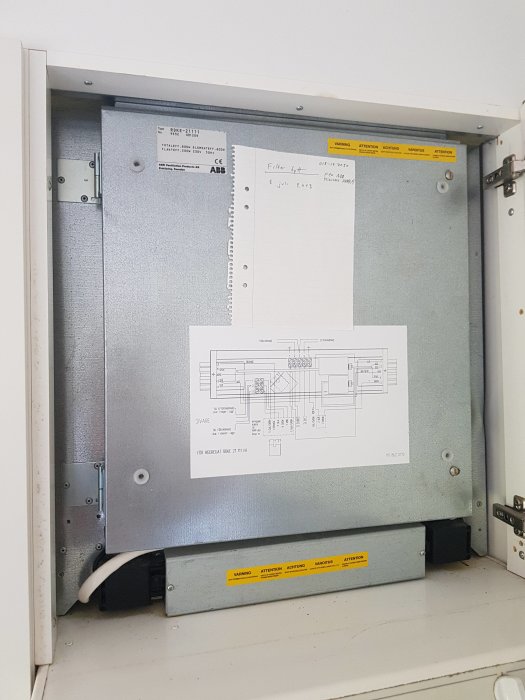 Mekanisk ventilationsenhet inbyggd bakom köksskåp med teknisk ritning och varningsetiketter.