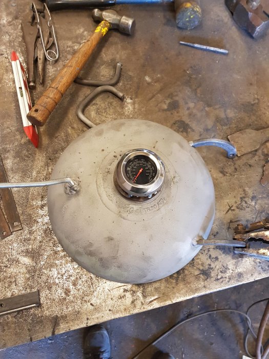 Arbetsbänk med verktyg och en grå rund anordning med mätare, eventuellt en del av en bil eller maskin.
