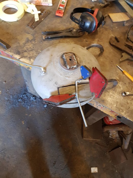 Gasflaska ligger på arbetsbänk med verktyg, svetshandskar och tvingar runtom, under svetsning eller reparation.
