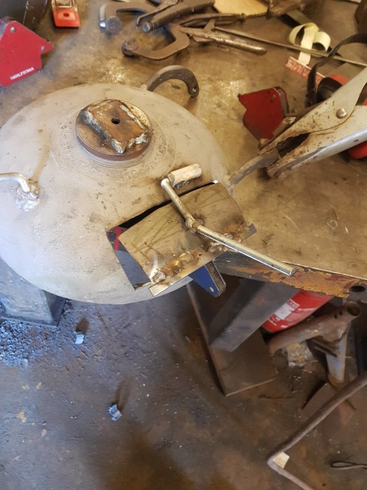 Svetstillbehör och verktyg på ett smutsigt arbetsbord med metallföremål i förgrunden.