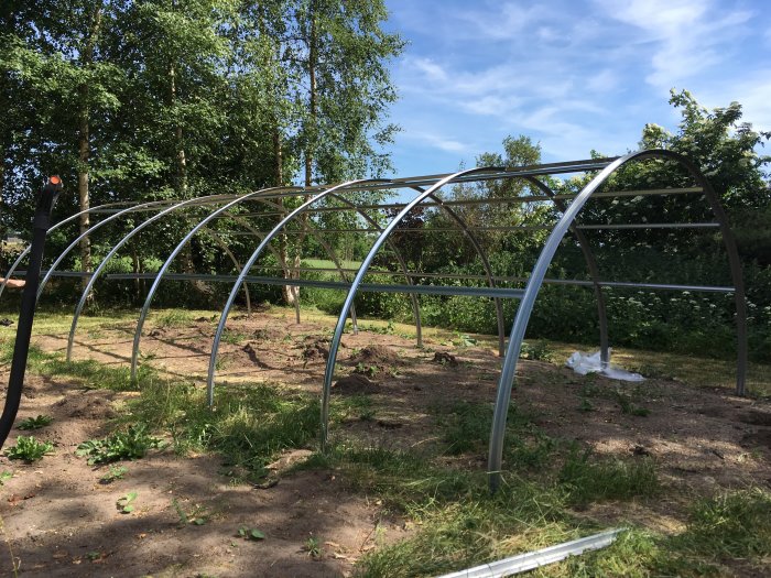 Halvfärdigt metallspindelskelett för byggprojekt på en gräsbevuxen tomt med träd i bakgrunden.