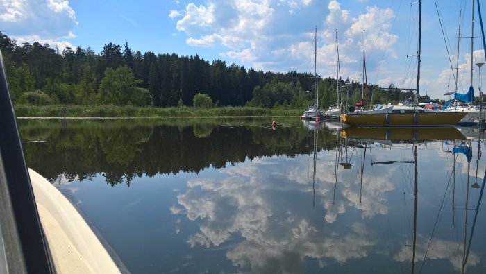 Idylliskt sommarlandskap med spegling av båtar och moln i lugnt sjövatten.