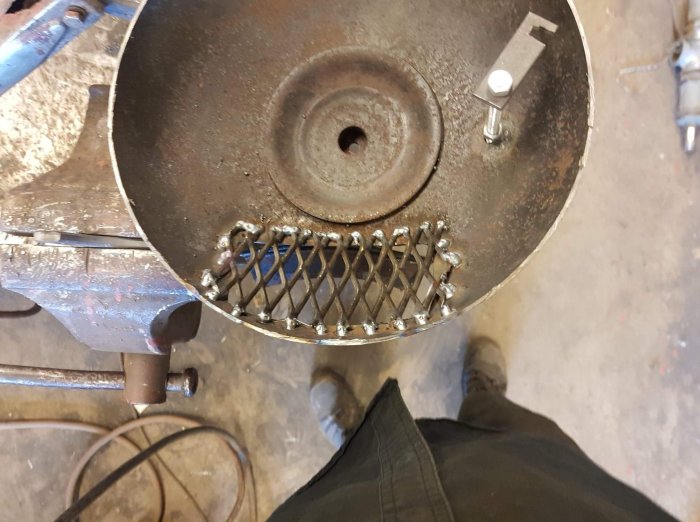 Svetsad metallgaller i öppningen av en ståltrumma, sett uppifrån med verktygsbord och personens skor i bakgrunden.