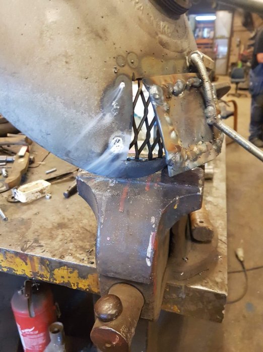 Svetsning av metallkonstruktion på en städ med synlig svetslåga och rök.